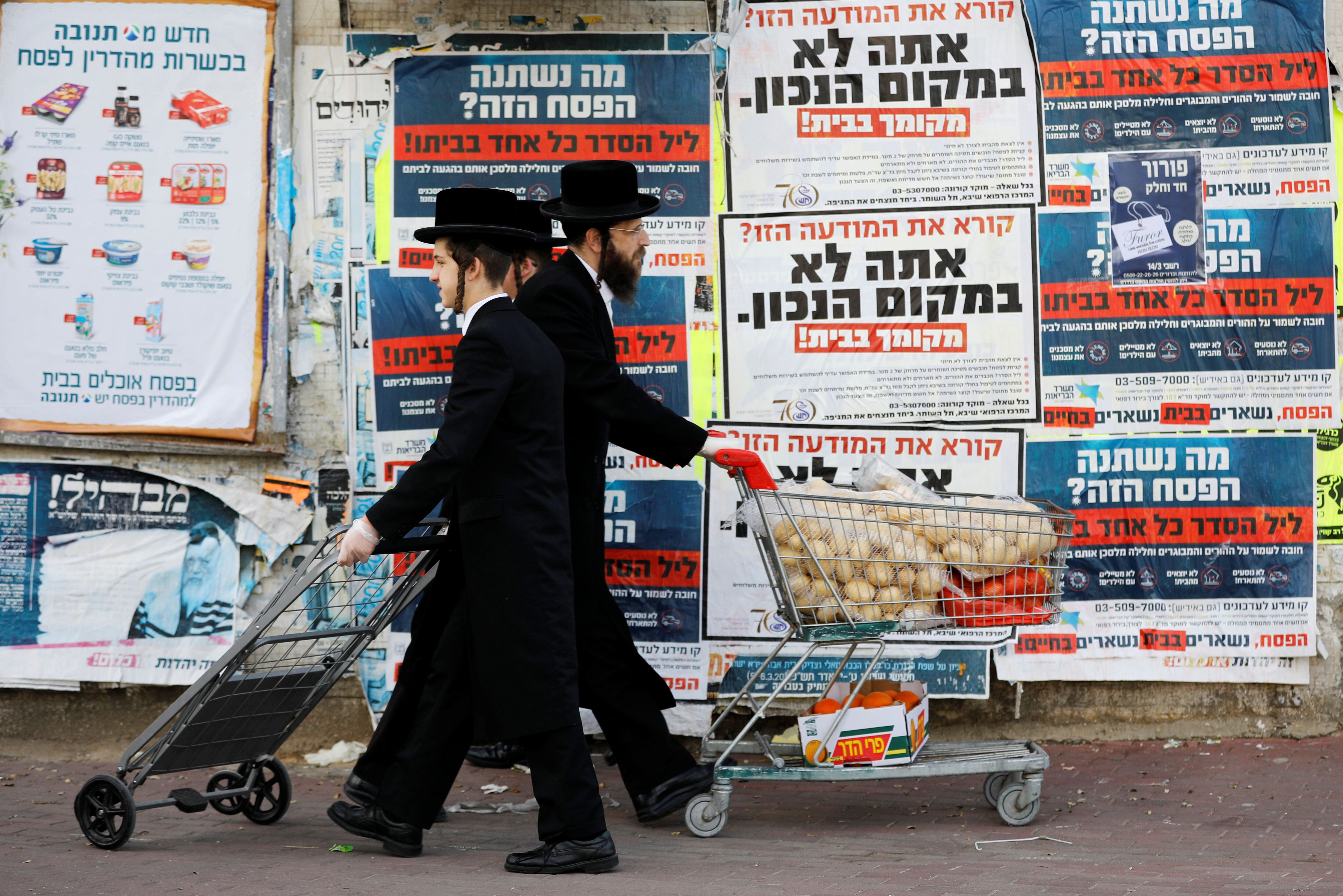 Judíos ultraortodoxos empujan carritos con comida frente a carteles que instan a la población a quedarse en sus casas por el coronavirus en Ashdod, Israel, el 1 de abril de 2020 (REUTERS/Amir Cohen)