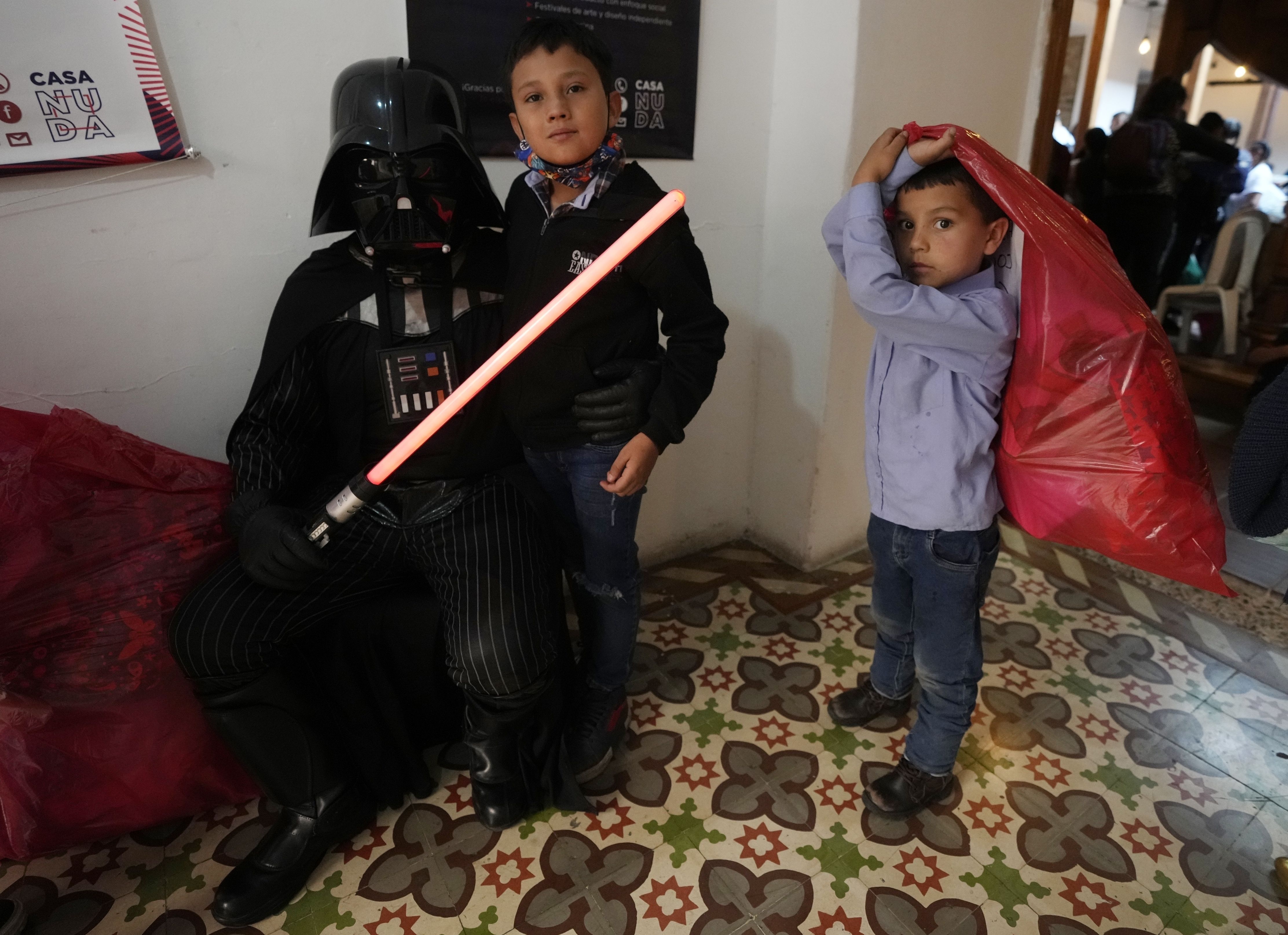 Un niño posa con un voluntario disfrazado de Darth Vader mientras otro niño sujeta una bolsa de regalo que recibió en un evento solidario organizado por la fundación "Vida Nueva" que trabaja para ayudar a extrabajadoras sexuales y víctimas de violencia, en Bogotá, Colombia, el jueves 22 de diciembre de 2022. (AP Foto/Fernando Vergara)